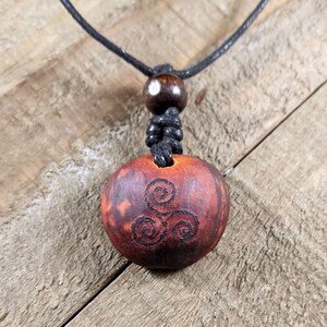 May include: A wooden pendant necklace with a dark brown, circular pendant. The pendant features a carved spiral design and is suspended from a black cord. The necklace includes a wooden bead and a knotted section.