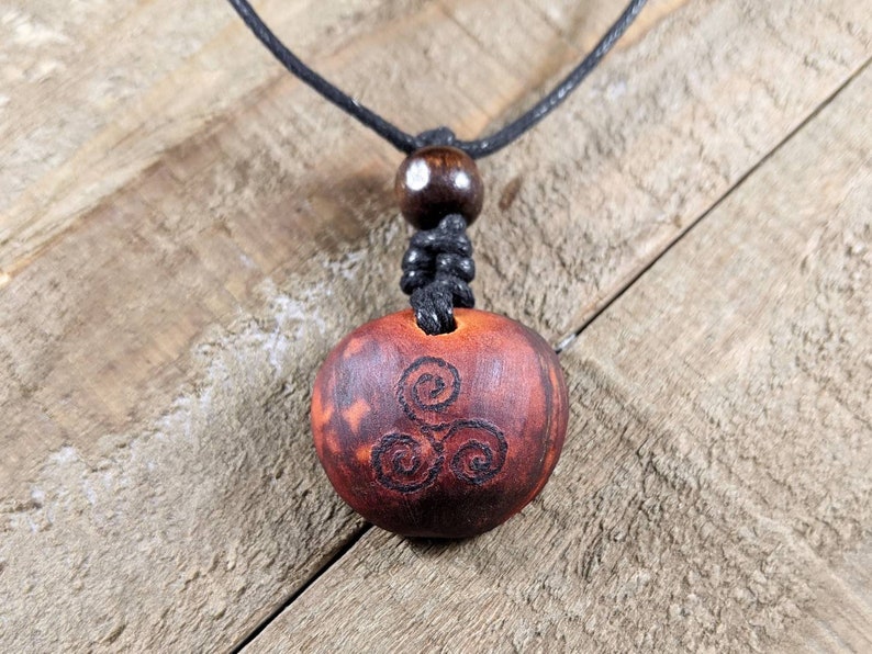May include: A wooden pendant necklace with a dark brown, circular pendant. The pendant features a carved spiral design and is suspended from a black cord. The necklace includes a wooden bead and a knotted section.