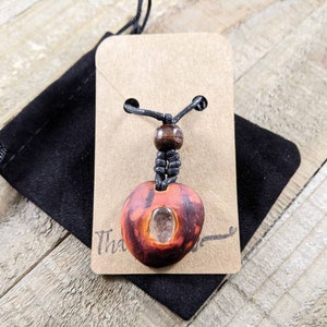 May include: A handmade pendant necklace featuring a reddish-brown, oval-shaped pendant with a clear stone insert. The pendant hangs from a black cord with a wooden bead accent. The necklace is displayed on a tan card with the word "Thank" visible.