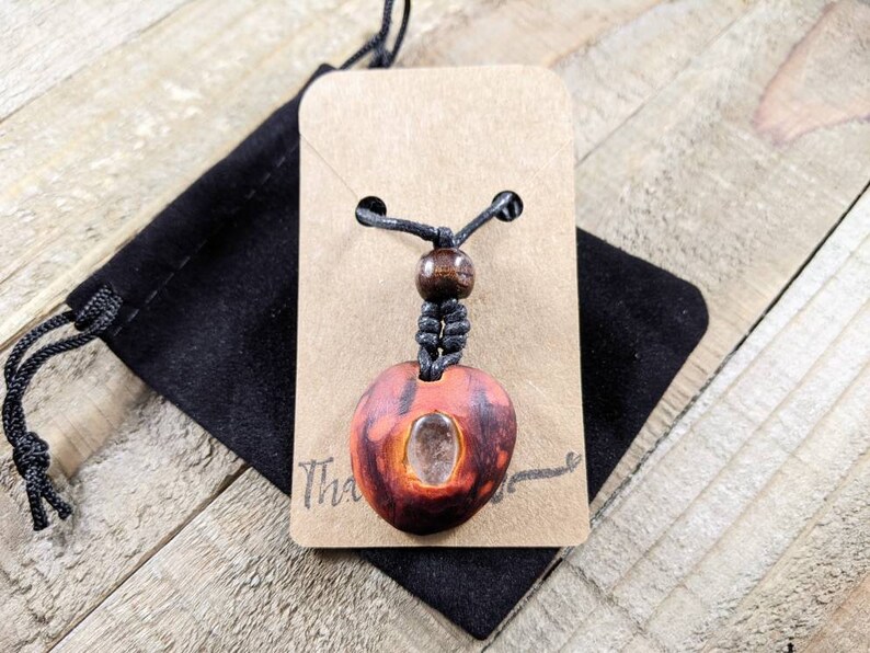 May include: A handmade pendant necklace featuring a reddish-brown, oval-shaped pendant with a clear stone insert. The pendant hangs from a black cord with a wooden bead accent. The necklace is displayed on a tan card with the word "Thank" visible.