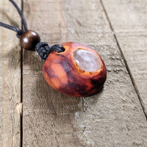 Pu&ograve; includere: Una collana con pendente fatta a mano con una pietra di forma organica marrone-rossastra con un centro chiaro e lucido. Il pendente &egrave; sospeso a un cordino nero con una perla di legno. La collana &egrave; esposta su una superficie di legno.