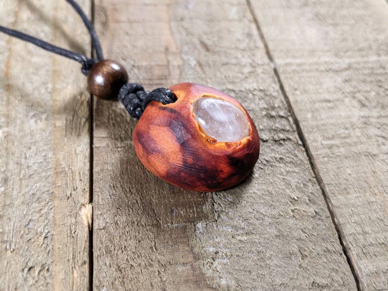 Pu&ograve; includere: Una collana con pendente fatta a mano con una pietra di forma organica marrone-rossastra con un centro chiaro e lucido. Il pendente &egrave; sospeso a un cordino nero con una perla di legno. La collana &egrave; esposta su una superficie di legno.