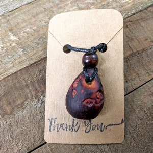 May include: A brown wooden pendant with intricate carvings hangs from a black cord with a brown wooden bead. The pendant is on a brown card with the text "Thank You".