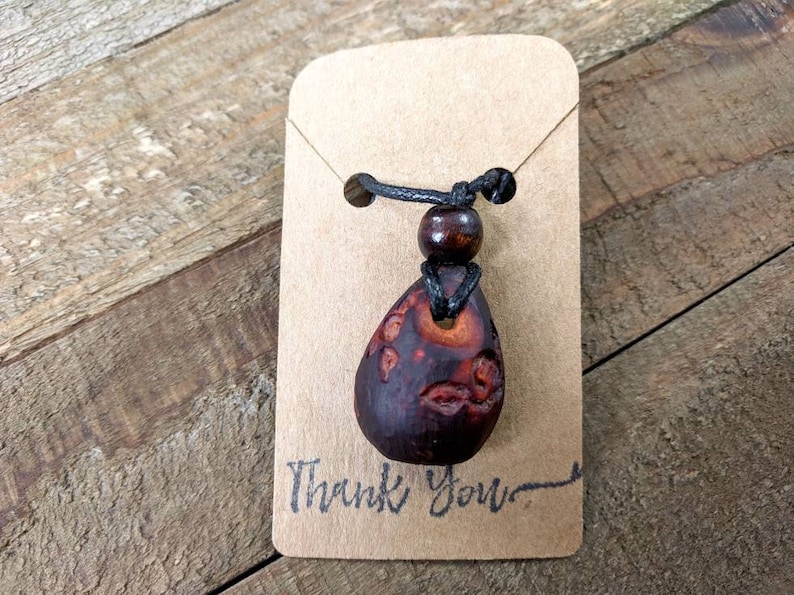 May include: A brown wooden pendant with intricate carvings hangs from a black cord with a brown wooden bead. The pendant is on a brown card with the text "Thank You".