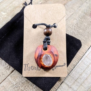 Pu&ograve; includere: Una collana con pendente in legno fatto a mano con un inserto di cristallo trasparente. Il pendente &egrave; di colore marrone-rossastro con venature naturali del legno. La collana &egrave; esposta su un cartoncino marrone con la scritta "Thank" e un sacchetto di velluto nero.