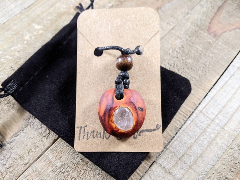 Pu&ograve; includere: Una collana con pendente in legno fatto a mano con un inserto di cristallo trasparente. Il pendente &egrave; di colore marrone-rossastro con venature naturali del legno. La collana &egrave; esposta su un cartoncino marrone con la scritta "Thank" e un sacchetto di velluto nero.