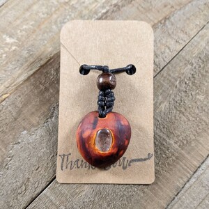 May include: A handmade wooden pendant necklace with a heart-shaped pendant. The pendant is reddish-brown with a clear stone insert. It hangs from a black cord with a wooden bead accent. The necklace is attached to a brown card that says "Thank you".