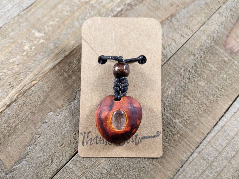 May include: A handmade wooden pendant necklace with a heart-shaped pendant. The pendant is reddish-brown with a clear stone insert. It hangs from a black cord with a wooden bead accent. The necklace is attached to a brown card that says "Thank you".