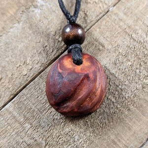May include: A handcrafted wooden pendant necklace. The pendant is a reddish-brown color with a carved spiral design. The necklace has a black cord and a wooden bead above the pendant.