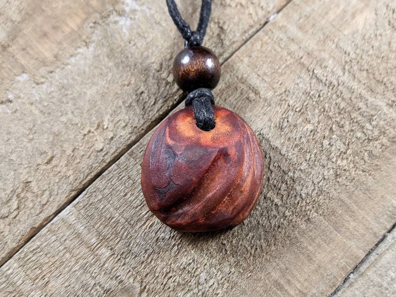 May include: A handcrafted wooden pendant necklace. The pendant is a reddish-brown color with a carved spiral design. The necklace has a black cord and a wooden bead above the pendant.