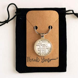 May include: A silver keychain with a round glass charm featuring a vintage dictionary definition of the word "traveler". The keychain is displayed on a brown card with the words "Thank You" written in black cursive. The keychain is packaged in a black velvet bag.