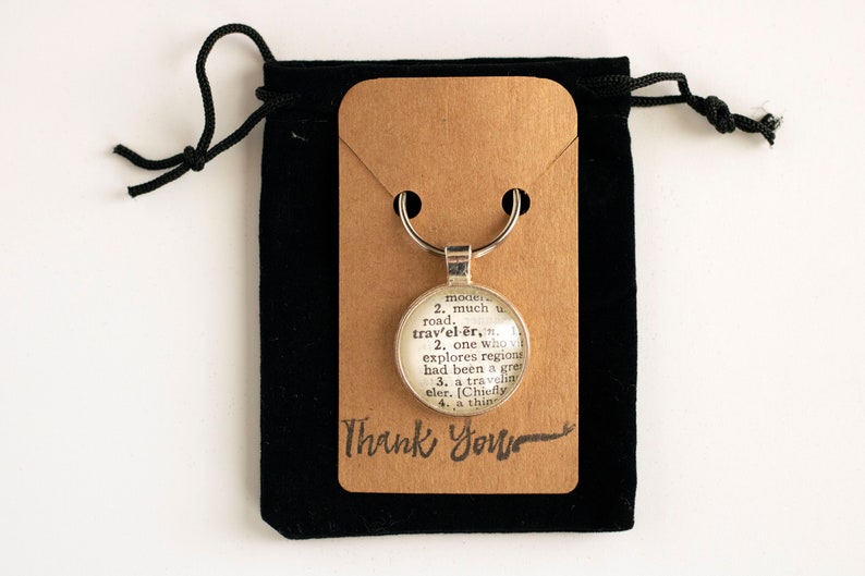 May include: A silver keychain with a round glass charm featuring a vintage dictionary definition of the word "traveler". The keychain is displayed on a brown card with the words "Thank You" written in black cursive. The keychain is packaged in a black velvet bag.