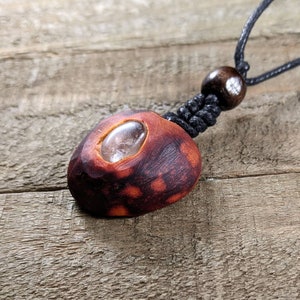 May include: A handcrafted pendant necklace featuring a reddish-brown wooden bead with a clear, oval-shaped stone inset. The pendant is suspended from a black cord with a wooden bead accent.