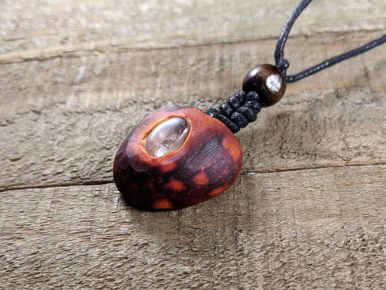 May include: A handcrafted pendant necklace featuring a reddish-brown wooden bead with a clear, oval-shaped stone inset. The pendant is suspended from a black cord with a wooden bead accent.