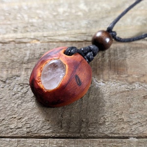 Pu&ograve; includere: Collana con pendente in legno fatto a mano. Il pendente &egrave; di colore marrone-rossastro con una pietra trasparente di forma ovale al centro. La collana ha un cordino nero con una perla di legno e un nodo scorrevole.