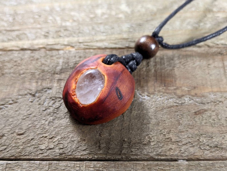 Pu&ograve; includere: Collana con pendente in legno fatto a mano. Il pendente &egrave; di colore marrone-rossastro con una pietra trasparente di forma ovale al centro. La collana ha un cordino nero con una perla di legno e un nodo scorrevole.