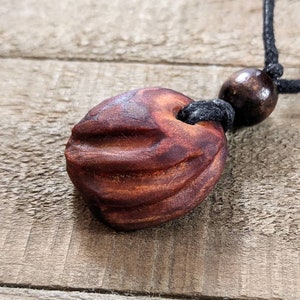 May include: A handcrafted wooden pendant necklace. The pendant is a reddish-brown, ridged, oval-shaped bead. It hangs from a black cord with a smaller, round wooden bead. The background is a weathered wooden surface.