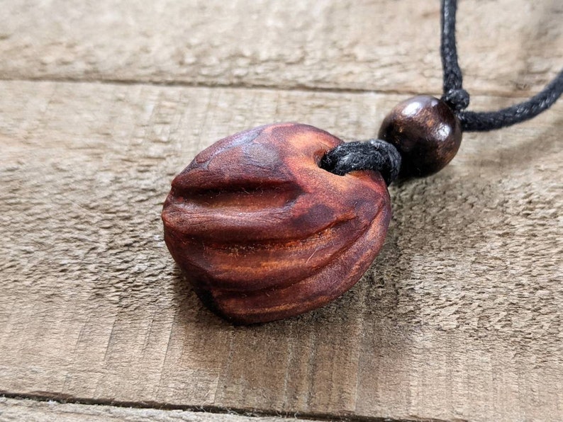 May include: A handcrafted wooden pendant necklace. The pendant is a reddish-brown, ridged, oval-shaped bead. It hangs from a black cord with a smaller, round wooden bead. The background is a weathered wooden surface.