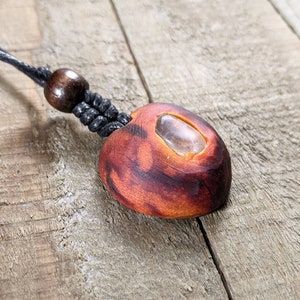 May include: A handmade pendant necklace featuring a reddish-brown, oval-shaped wooden pendant with a clear stone inlay. The pendant is suspended from a black cord with a wooden bead accent. The necklace is displayed on a wooden surface.