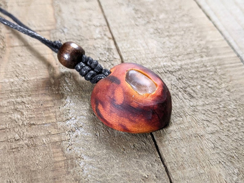 May include: A handmade pendant necklace featuring a reddish-brown, oval-shaped wooden pendant with a clear stone inlay. The pendant is suspended from a black cord with a wooden bead accent. The necklace is displayed on a wooden surface.