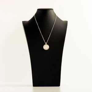 May include: A silver chain necklace with a round, gold pendant featuring a white, sparkly design. The necklace is displayed on a black mannequin bust.