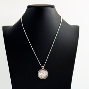 Puede incluir: Un collar de cadena de plata con un colgante redondo con impresi&oacute;n de texto en blanco y negro. El colgante est&aacute; en un expositor de joyer&iacute;a negro.