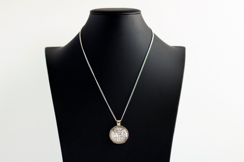 Puede incluir: Un collar de cadena de plata con un colgante redondo con impresi&oacute;n de texto en blanco y negro. El colgante est&aacute; en un expositor de joyer&iacute;a negro.