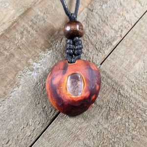 May include: A handcrafted pendant necklace featuring a reddish-brown, heart-shaped wooden pendant with a clear, oval-shaped crystal embedded in the center. The necklace has a black cord and a wooden bead.