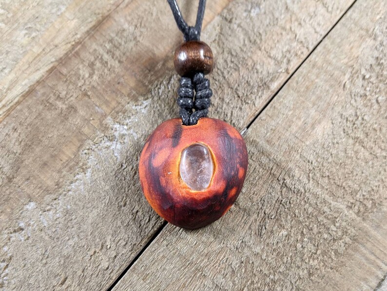 May include: A handcrafted pendant necklace featuring a reddish-brown, heart-shaped wooden pendant with a clear, oval-shaped crystal embedded in the center. The necklace has a black cord and a wooden bead.