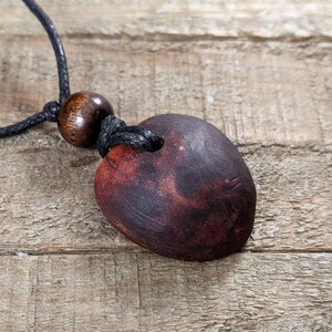 May include: A handmade wooden pendant necklace. The pendant is a dark brown, heart-shaped piece with a hole at the top, strung on a black cord. A round wooden bead is also on the cord. The necklace rests on a wooden surface.