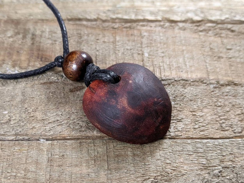 May include: A handmade wooden pendant necklace. The pendant is a dark brown, heart-shaped piece with a hole at the top, strung on a black cord. A round wooden bead is also on the cord. The necklace rests on a wooden surface.