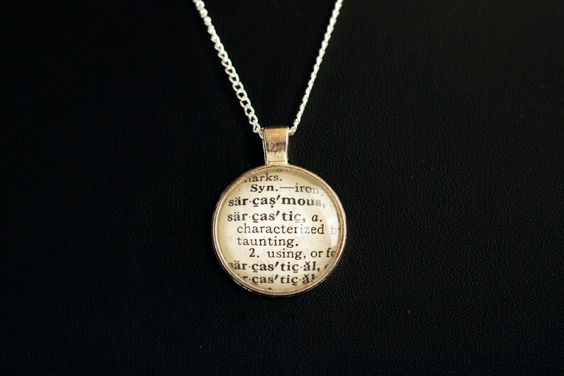 May include: A silver necklace with a round pendant featuring a dictionary definition of the word "sarcastic". The pendant is made of glass and has a vintage look.