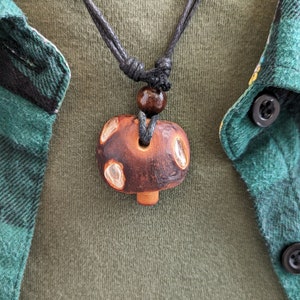 May include: A brown wooden mushroom pendant necklace with a black cord and a brown wooden bead. The necklace is worn by a person wearing a green and black plaid shirt.