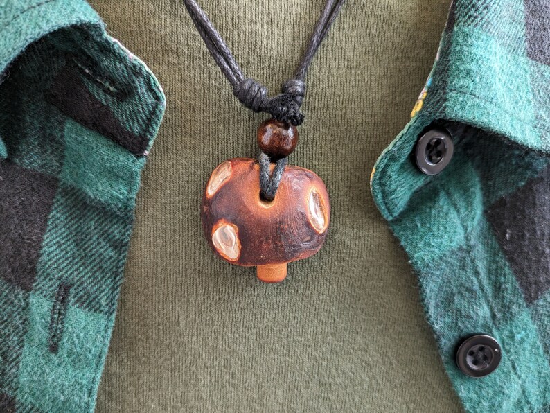 May include: A brown wooden mushroom pendant necklace with a black cord and a brown wooden bead. The necklace is worn by a person wearing a green and black plaid shirt.