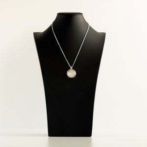 Puede incluir: Un collar de cadena de plata con un colgante redondo que presenta un dise&ntilde;o detallado. El collar se muestra en un busto de maniqu&iacute; negro.
