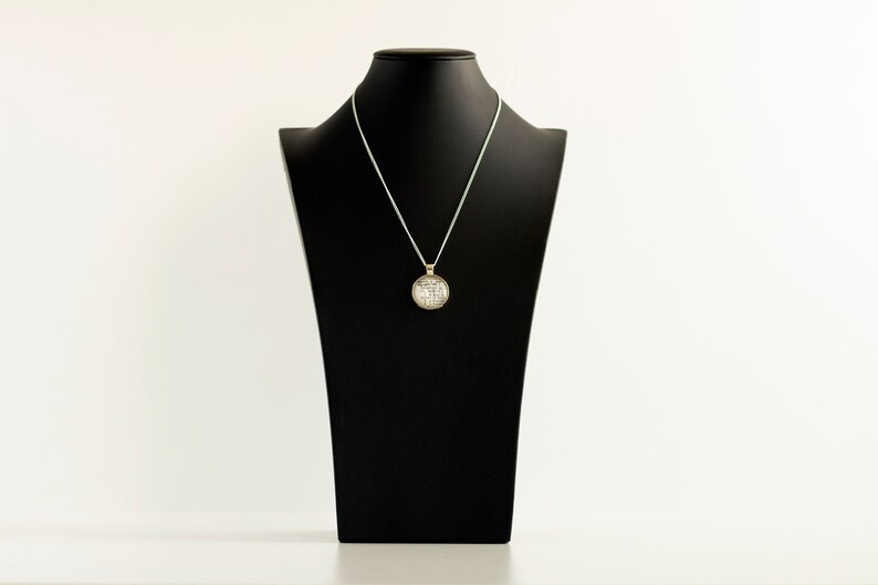Puede incluir: Un collar de cadena de plata con un colgante redondo que presenta un dise&ntilde;o detallado. El collar se muestra en un busto de maniqu&iacute; negro.