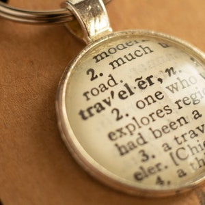 May include: A silver keychain with a round glass charm. The charm features a vintage dictionary definition of the word "traveler". The definition reads: "2. one who explores regions, had been a great traveler. [Chiefly a thing of the past]"