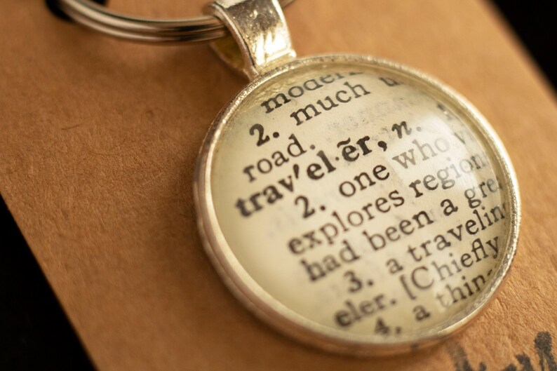 May include: A silver keychain with a round glass charm. The charm features a vintage dictionary definition of the word "traveler". The definition reads: "2. one who explores regions, had been a great traveler. [Chiefly a thing of the past]"