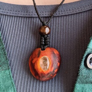 May include: A handmade wooden pendant necklace. The pendant is heart-shaped with a clear stone inlay and a dark brown finish. The necklace has a black cord and a wooden bead. The person is wearing a blue ribbed shirt and a green and black plaid shirt.
