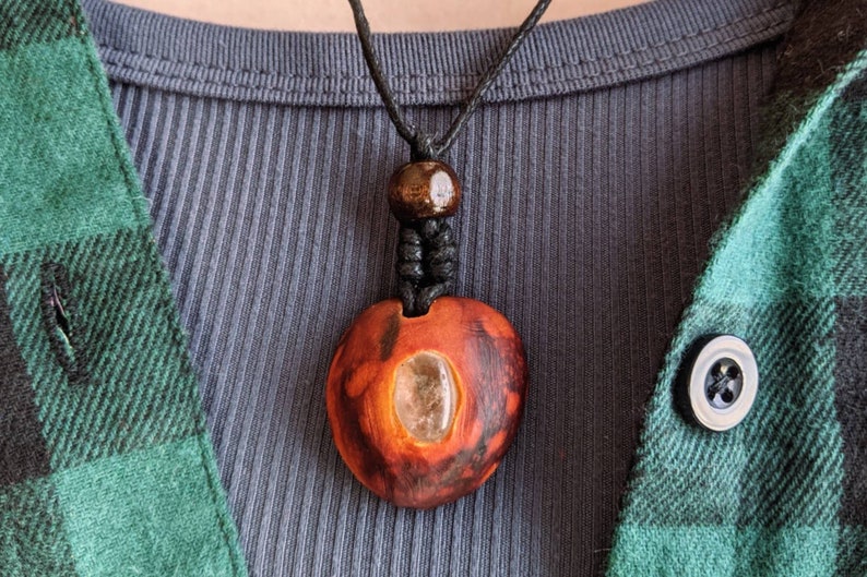May include: A handmade wooden pendant necklace. The pendant is heart-shaped with a clear stone inlay and a dark brown finish. The necklace has a black cord and a wooden bead. The person is wearing a blue ribbed shirt and a green and black plaid shirt.