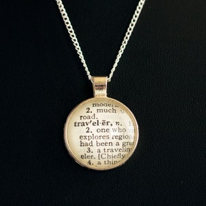May include: A silver chain necklace with a round pendant featuring a vintage dictionary definition of the word "traveler".