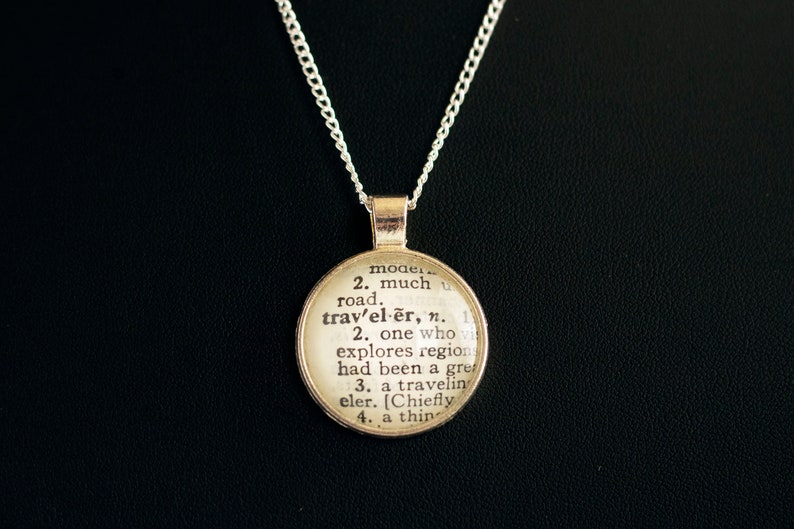 May include: A silver chain necklace with a round pendant featuring a vintage dictionary definition of the word "traveler".