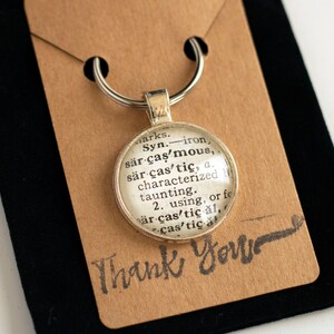 May include: A silver keychain with a round glass charm featuring a dictionary definition of the word "sarcastic". The charm is attached to a silver key ring. The background is a brown paper tag with the words "Thank You" written in black.