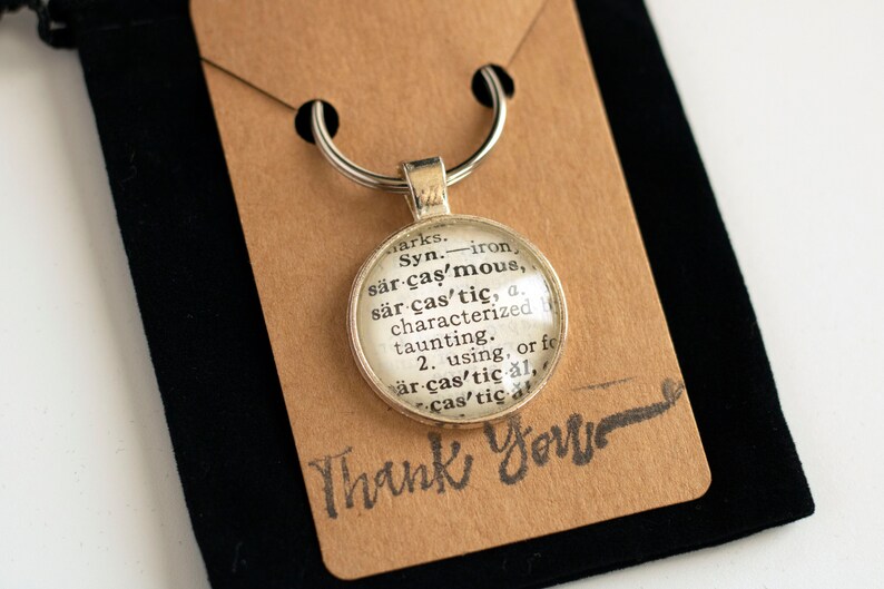 May include: A silver keychain with a round glass charm featuring a dictionary definition of the word "sarcastic". The charm is attached to a silver key ring. The background is a brown paper tag with the words "Thank You" written in black.