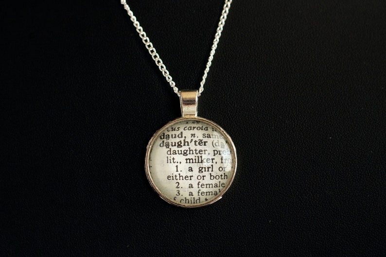 Puede incluir: Un collar de cadena de plata con un colgante redondo que presenta una definici&oacute;n del diccionario de la palabra "hija".