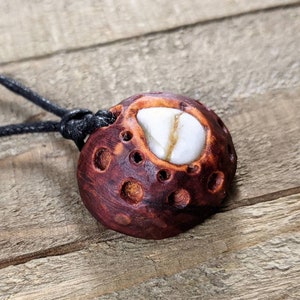 May include: A dark brown wooden pendant with a white stone inlay, resembling a moon with craters. The pendant is attached to a black cord.