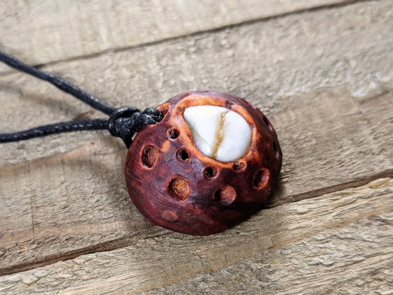 May include: A dark brown wooden pendant with a white stone inlay, resembling a moon with craters. The pendant is attached to a black cord.
