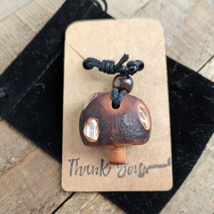 May include: A brown ceramic mushroom pendant with white accents hangs from a black cord with a brown wooden bead. The pendant is displayed on a brown card with the text "Thank You" written on it.