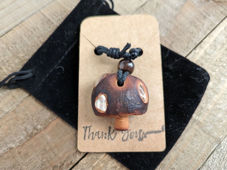 May include: A brown ceramic mushroom pendant with white accents hangs from a black cord with a brown wooden bead. The pendant is displayed on a brown card with the text "Thank You" written on it.