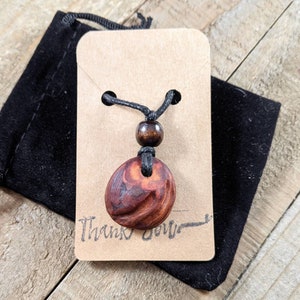 May include: A wooden pendant necklace with a dark brown, oval-shaped pendant. The pendant hangs from a black cord with a wooden bead. The necklace is displayed on a brown tag with the words "Thank you" and a black velvet pouch.